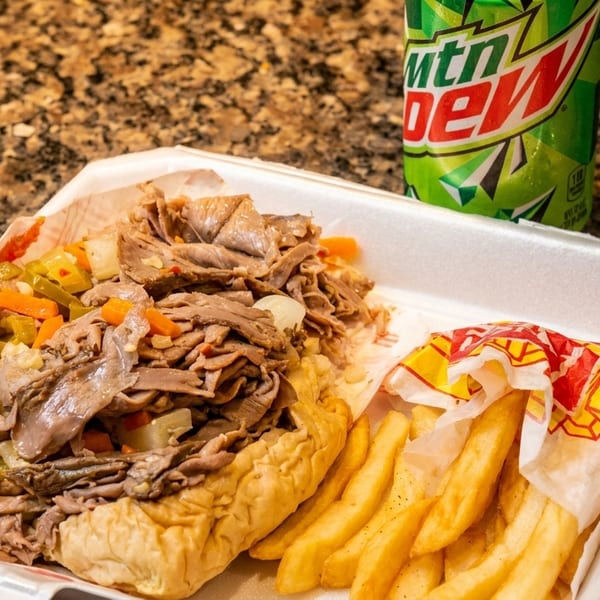 Italian beef sandwich with giardiniera, fries, a side of sauce, and a Mountain Dew can.